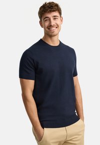 Granatowy sweter z krótkim rękawem, dzianinowy, z okrągłym dekoltem, prążkowanymi mankietami i dolnym obszyciem. Materiał wydaje się miękki o gładkiej fakturze.