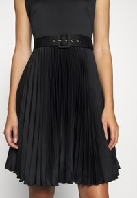 Svart plisserad klänning med figursydd överdel och ett brett tygbälte med fyrkantigt spänne. Materialet verkar mjukt med en lätt glans.