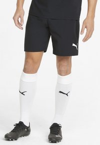 Mustard sportlikud lühikesed püksid valge Puma logoga, koos põlve kõrguste valgete sokkidega, millel on must logo, ja mustade jalgpallikleitidega, millel on tekstuuriline disain.