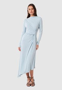 Light blue long-sleeve dress in a soft, smooth fabric with a twisted wrap design at the waist and an asymmetrical hem. Black strappy sandals.