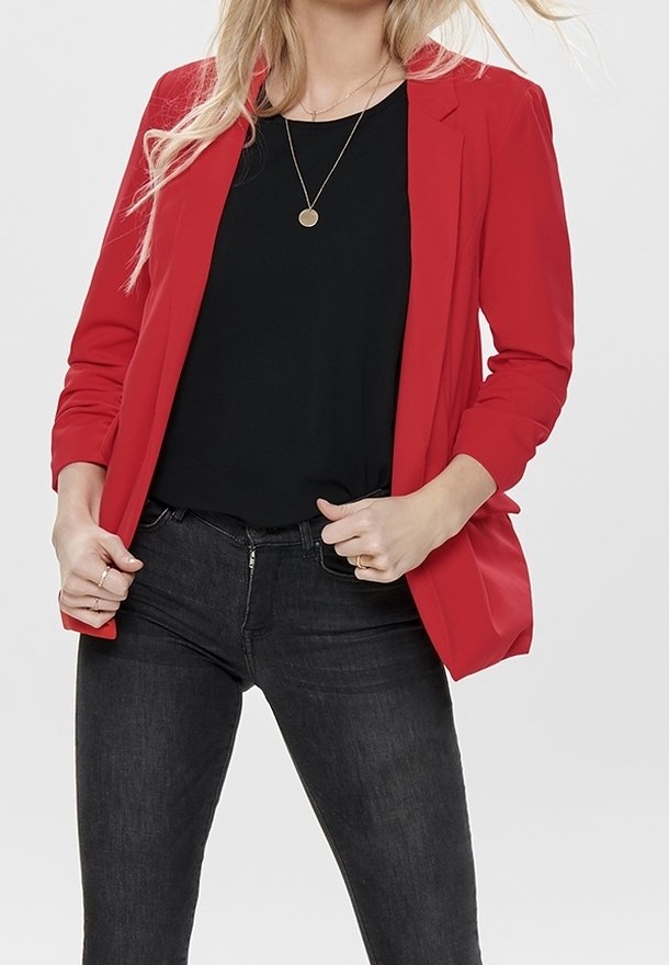 Blazer rojo con un corte ajustado, mangas largas y solapas con muesca, combinado con una blusa negra y jeans oscuros, creando un look en capas.