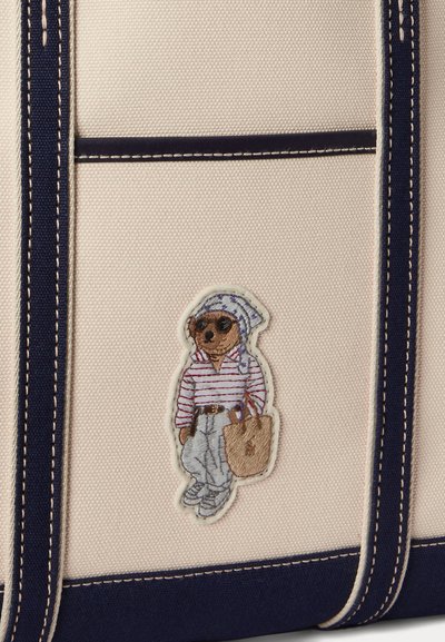 Beige and navy tote bag with an embroidered bear wearing sunglasses, a striped shirt, pants, a headscarf, and holding a tote bag.