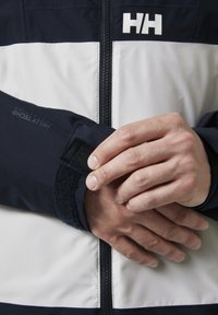 Marineblauwe en witte waterdichte jas met een ritsluiting, voorzien van een klittenband manchet en logodetail, afgebeeld op een hand die de mouw aanpast.