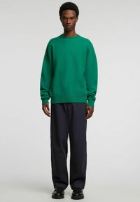 Zielony sweter na drutach z okrągłym dekoltem, długimi rękawami i ściągaczem u dołu, połączony z ciemnoniebieskimi szerokimi spodniami i czarnymi butami.