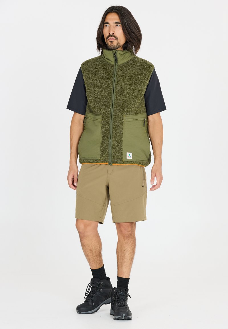 Olivgrön fleeceväst med dragkedja och två framfickor, tillsammans med beige shorts och svarta vandrarskor. Enkel svart t-shirt med kort ärm under.