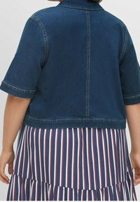 Jeansjacka med korta ärmar, mörkblått tyg, synliga sömmar och en något kortare design, bärs över en randig klänning med röda och blå ränder.