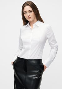 Γυναίκα με μακριά καστανά μαλλιά, φορώντας φορεμένη λευκή πουκαμίσα με κουμπιά και μαύρη δερμάτινη φούστα, στέκεται με τα χέρια στις τσέπες.