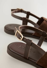 Braune Leder-Sandalen mit mehreren Riemen, einer goldenen Schnalle, einem weichen Wildlederelement und einer flachen, strukturierten Sohle.