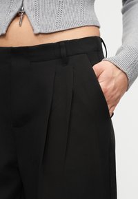 Κοντινό πλάνο ενός ατόμου που φοράει μαύρο πλισέ παντελόνι με το χέρι στην τσέπη και ένα κοντό γκρι πλεκτό ζακέτα με φερμουάρ.