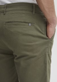 Olivenfargede bomullsbukser med en baklomme med svart-hvitt stripet dekor og klassisk rett ben-design.