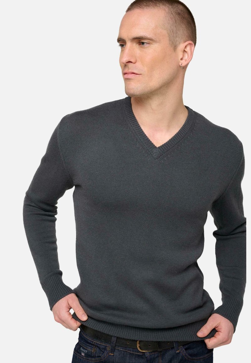 Pull en v gris à côtes avec des manches longues. Il présente une finition texturée et une silhouette ajustée, associé à un jean en denim foncé.