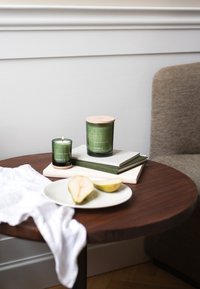 Green glass candles with wooden lids are placed on stacked books atop a round wooden table. A white plate holds pear slices.