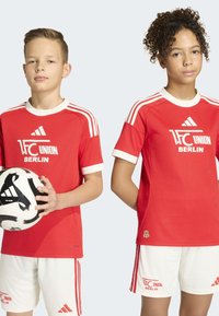 Deux enfants portant des maillots rouges du 1. FC Union Berlin et des shorts blancs, dont l'un tient un ballon de football noir et blanc.