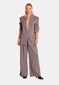 Blazer gris à rayures, avec épaules rembourrées, décolleté en V profond et pantalon large assorti, présentant un tissu texturé et des accents brillants.