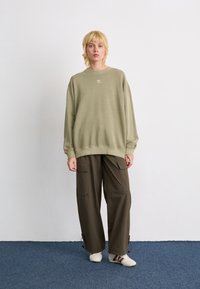 Person med kort blond hår iført en overdimensioneret olivengrøn sweatshirt, mørkebrune cargo-bukser og hvide sneakers, der står på et blåt tæppe.