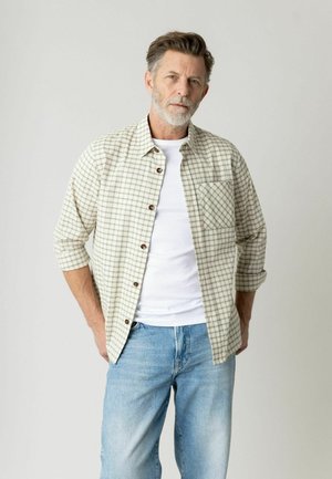 Mellemaldrene mand med gråt hår og skæg iført hvid t-shirt, beige ternet skjorte og lyseblå jeans, stående med hænderne i lommerne.