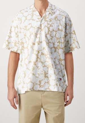 Man wearing short-sleeve button-up shirt with beige and white floral pattern and beige pants, standing against plain background.