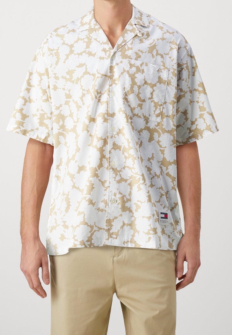 Homme portant une chemise à manches courtes à boutons avec un motif floral beige et blanc et un pantalon beige, debout devant un fond uni.