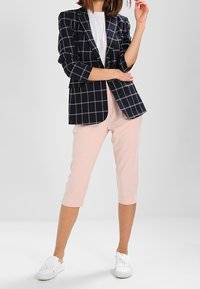 Blazer à carreaux bleu marine avec des lignes blanches et grises, porté sur une chemise blanche. Un pantalon court rose clair et des chaussures blanches complètent la tenue.
