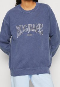 Blå sweatshirt i mjukt tyg, med vit broderad text "BDG JEANS" i en rundad design, samt ribbstickade muddar och krage.
