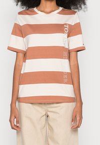 Camiseta de rayas con colores alternos terracota y crema. Fabricada en algodón, con mangas cortas y escote redondo. Detalle de logo en el lateral.