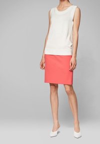 Un haut en tricot sans manches blanc associé à une jupe crayon corail. Le modèle porte des chaussures blanches à bout pointu. Textures lisses, design minimaliste.