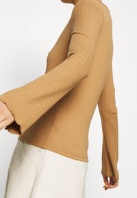 Chemise à manches longues côtelée de couleur brun clair, avec une coupe ajustée et des poignets évasés. Fabriquée en tissu extensible, elle présente une texture lisse et un léger mouvement.