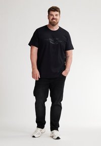 Camiseta negra con un diseño gráfico impreso, combinada con jeans negros y zapatillas deportivas blancas. Ajuste sencillo y casual, material de algodón suave.