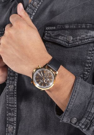 Montre analogique dorée avec bracelet en cuir noir, dotée d'un cadran gris foncé, d'accents argentés et de deux sous-cadrans ; portée au poignet.