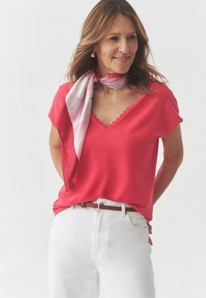 Raudona trumpomis rankovėmis palaidinė su V formos iškirpte ir banguota apdaila, derinama su baltais keliais ir šviesiai rožine raštuota šaliku aplink kaklą.