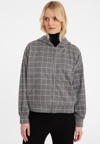 Haut à capuche zippé gris avec un motif en grille blanche. Tissu doux, coupe décontractée, manches longues avec poignets et une capuche. Conçu avec des poches latérales.