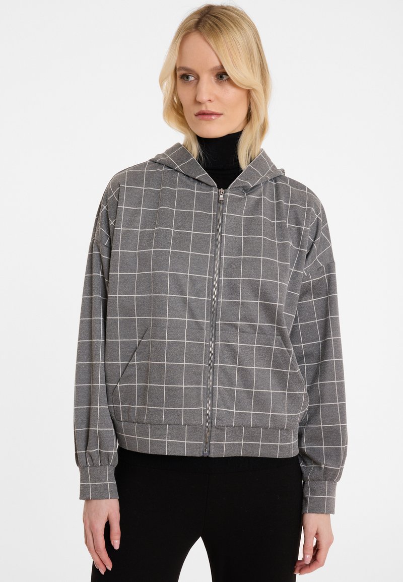 Haut à capuche zippé gris avec un motif en grille blanche. Tissu doux, coupe décontractée, manches longues avec poignets et une capuche. Conçu avec des poches latérales.