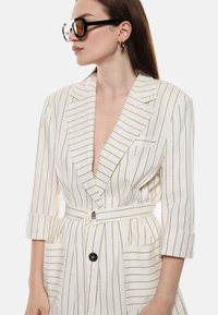 Abito blazer in lino off-white con righe blu navy, scollatura a V profonda, maniche arrotolate, una cintura con fibbia circolare dorata e tasche frontali.