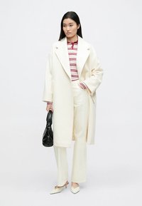 Valkoinen ylisuurikokoinen takki raidallisen punavalkoisen topin päällä, yhdistetty valkoisiin housuihin ja teräväkärkisiin korkokenkiin. Musta laukku kädessä.