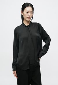 Čierna blúzka s dlhým rukávom z hladkej látky, s viazankou na krku a ozdobnými strapcami, s prehnutým lemom a manžetami na gombíky.