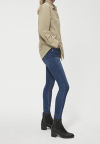 Chemise beige à boutons, jean bleu ajusté et bottines noires à talon épais. La tenue présente des textures lisses et une coupe sur mesure.