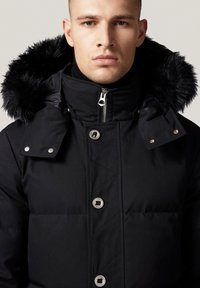 Czarna kurtka puchowa z wysokim kołnierzem, futrzanym kapturkiem, metalowymi elementami oraz srebrnymi zatrzaskami. Posiada pikowaną teksturę i elegancki design.