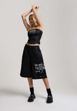 Donna con trecce lunghe che indossa un top nero senza spalline e pantaloncini neri larghi con grafica bianca, posando con le braccia alzate su sfondo grigio.