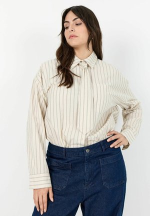 Woman wearing a cream-colored, striped blouse with a necktie and dark blue high-waisted jeans, standing with one hand on her hip.