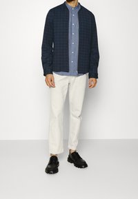 Uomo che indossa una giacca a zip a quadri blu navy, una camicia azzurra, pantaloni avorio e scarpe nere con lacci, in piedi contro uno sfondo semplice.