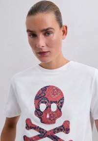 Biała bawełniana koszulka z kolorowym nadrukiem czaszki i skrzyżowanych kości, zdobionym skomplikowanymi wzorami w odcieniach czerwieni, niebieskiego i kremowego.