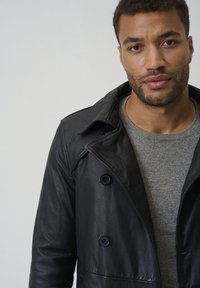 Zwart leren trenchcoat met een dubbele rij knopen en grote knopen. Gedragen over een grijze trui, met een spread collar en een gladde textuur.