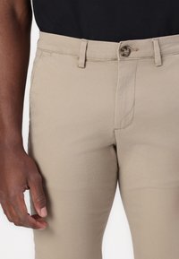 Beige chinos med slim fit, tillverkade av bomullsmix. Har en knappstängning och bälteshällor, med en jämn och mjuk yta.