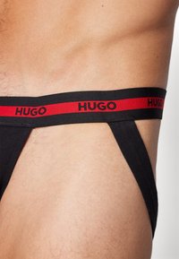 Schwarze Strings mit einem breiten Taillenbund und roter "HUGO"-Beschriftung, elastisches Material mit glatter Textur, minimalistisches Design.