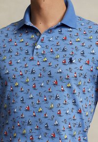 Blå polotröja med mönster av färgglada segelbåtar och ankor, med krage och knappar, som bärs av en person från nacken till mitten av torso.