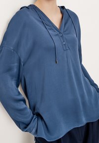 Pull bleu à manches longues avec encolure en V et capuche, comportant deux boutons et un cordon de serrage. Texture lisse avec une coupe décontractée.