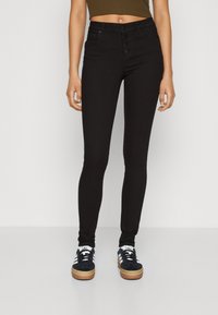 Svarte høytalende skinnyjeans med knappelukking, fem lommer og en glatt tekstur, kombinert med mørke joggesko.