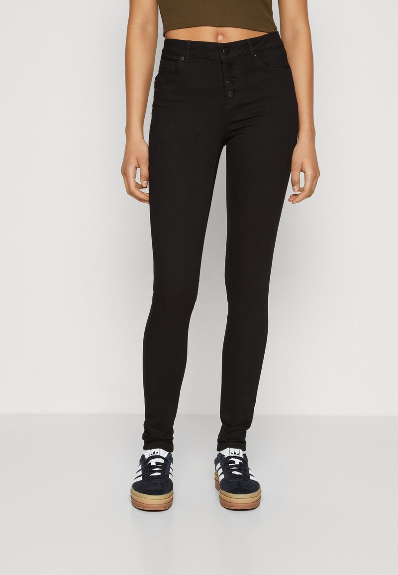 Svarte høytalende skinnyjeans med knappelukking, fem lommer og en glatt tekstur, kombinert med mørke joggesko.