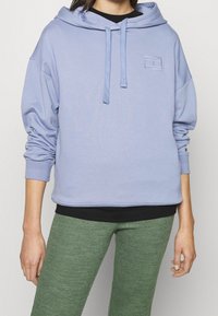 Sudadera azul claro con cordón, bolsillo frontal y pequeño logo bordado. Combinada con una parte superior negra oscura y leggings verdes texturizados.