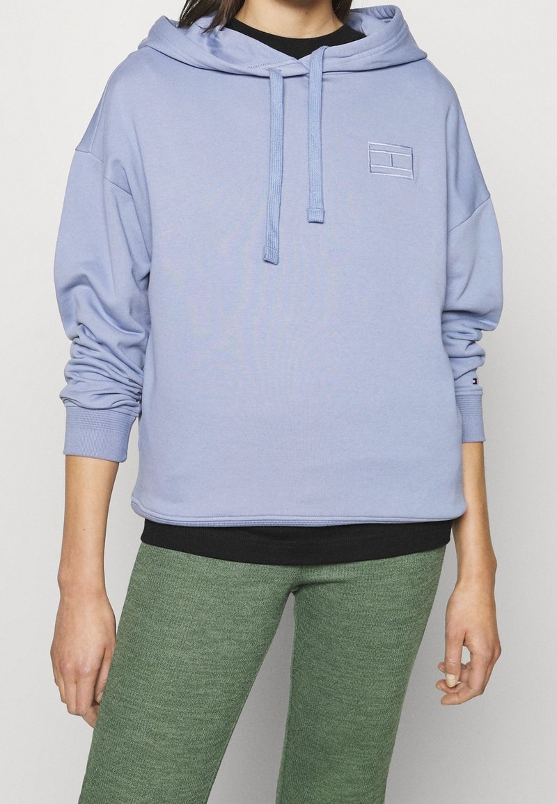 Sudadera azul claro con cordón, bolsillo frontal y pequeño logo bordado. Combinada con una parte superior negra oscura y leggings verdes texturizados.
